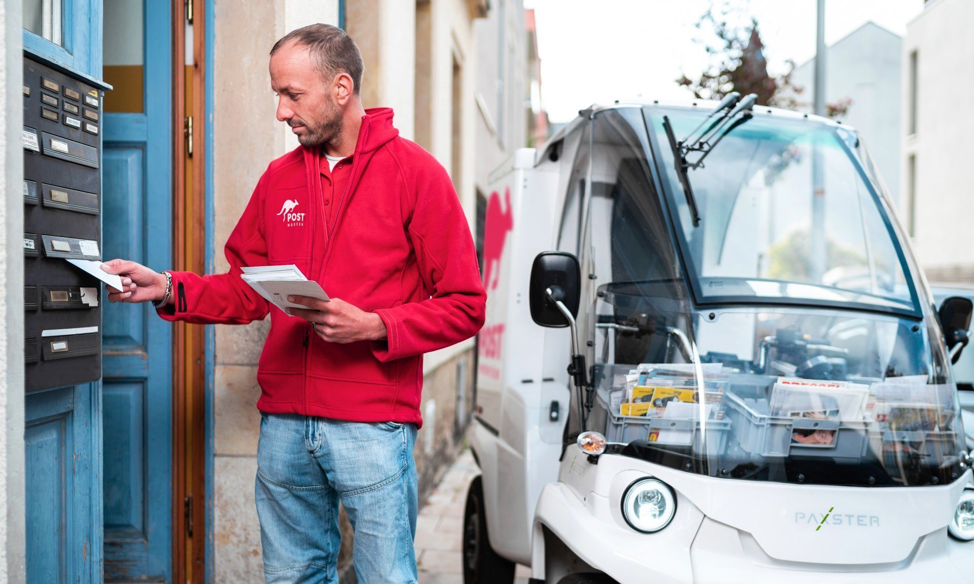Das Bild zeigt einen PostModern-Mitarbeiter mit einem Paxster, welcher gerade Post zustellt.