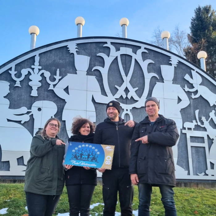 Das Bild zeigt den großen Schwibbogen in Johanngeorgenstadt. Davor stehen: v.l.n.r.: Susan Lamla, Kundenberaterin PostModern/ MEDIA Logistik Aue; Jana Busch, Hauptamtsleiterin Johanngeorgenstadt; Toni Bachmann, stellvertretender Bürgermeister Johanngeorgenstadt; Harald Teller, Nachfahre der Johanngeorgenstädter Schwibbogenschöpfer mit der Schaubriefmarke zur Sonderbriefmarke in der Wertstufe 80 Cent.